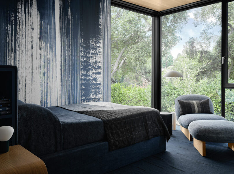 A modern bedroom with a blue-themed interior features a bed, cushioned chair, and floor lamp by large windows overlooking greenery.