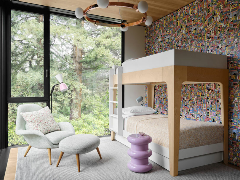 A modern bedroom with a large window, bunk bed, patterned wallpaper, cushioned chair with ottoman, pink side table, and a round chandelier.