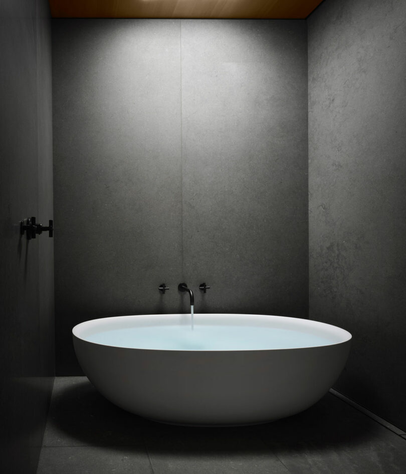 Minimalist bathroom with a cylindrical white bathtub filled with water, against dark gray walls and a wooden ceiling. A simple black faucet is mounted above the tub.