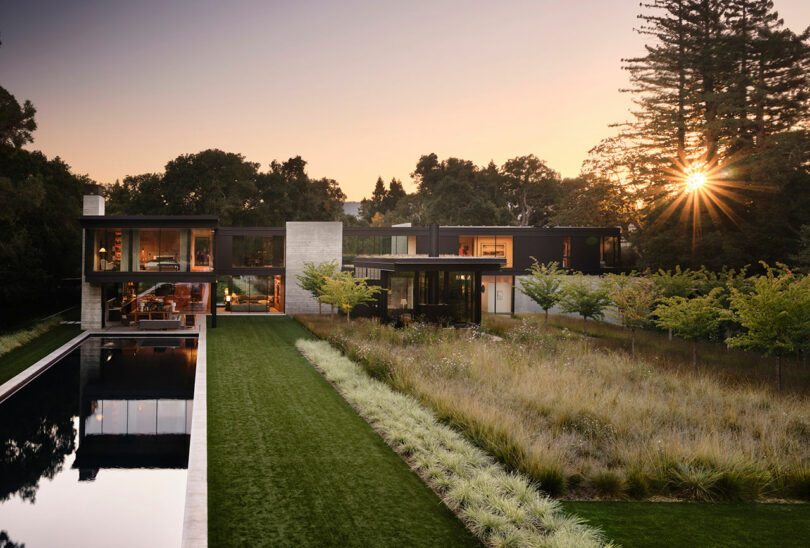 Modern house with large glass windows, surrounded by trees, a long pool on the left, and a landscaped garden. The sun sets in the background.