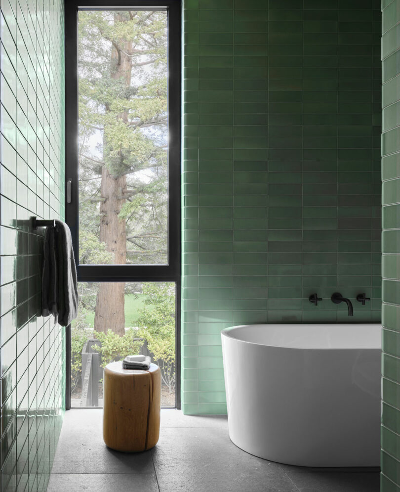 Modern bathroom with green tiled walls, a white bathtub, and a wooden stool next to a large window showing trees outside.