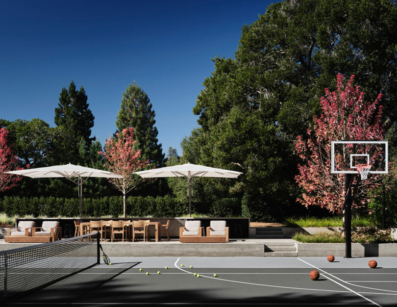 Outdoor sports court with tennis and basketball setups, surrounded by trees. There are seating areas with umbrellas nearby.