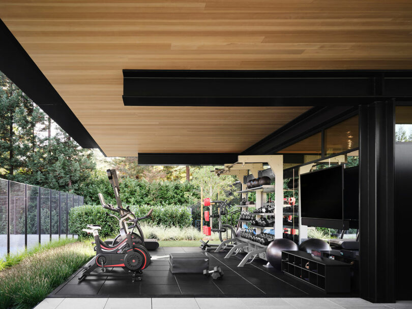 Modern outdoor home gym with exercise bike, treadmill, weights, and a punching bag on black flooring. Surrounded by greenery, the space has a large TV and a wooden ceiling.