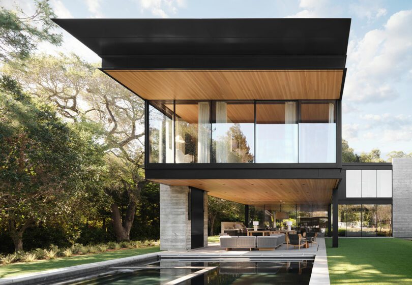 Modern house with a cantilevered upper floor featuring large glass windows, a pool in the foreground, and surrounded by trees.