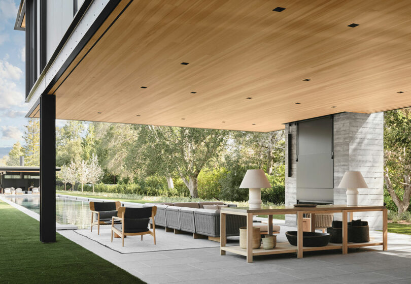 Modern outdoor patio with a wooden ceiling, comfortable seating, tables, and decorative lamps, overlooking a swimming pool and surrounded by greenery.