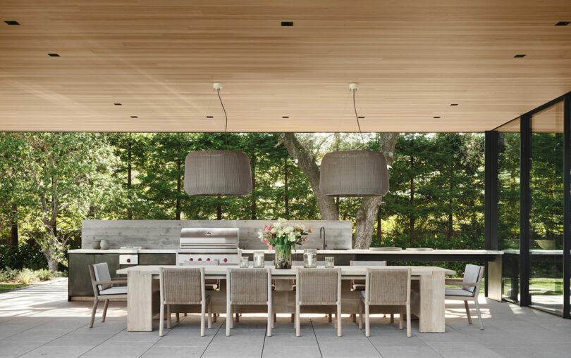 Modern outdoor kitchen with a long dining table set for eight, two large pendant lights, sleek countertops, and a barbecue grill, surrounded by a lush green forest.