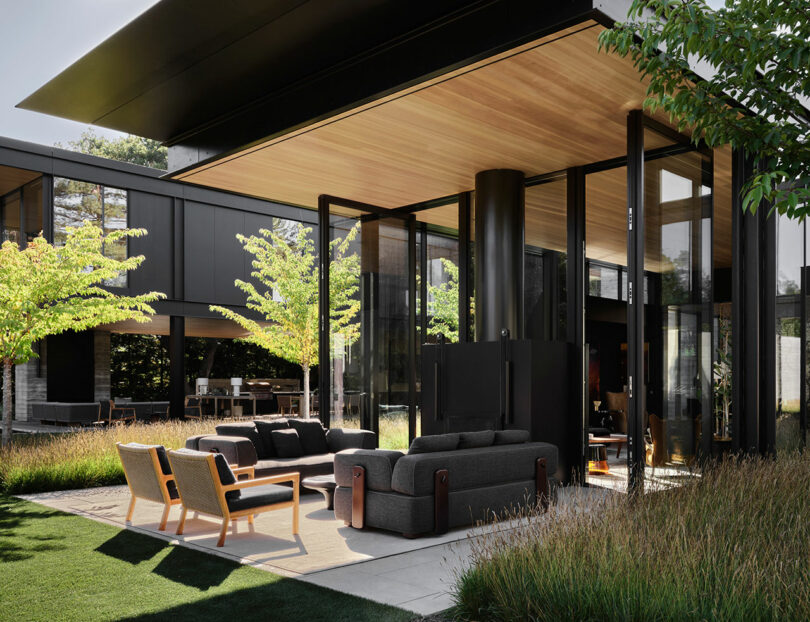 Modern outdoor patio with black framed glass walls, wooden ceiling, gray sofas, and chairs. Surrounded by grass and trees, the space extends from a contemporary building.