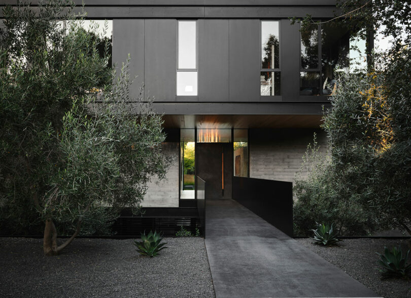 Modern black and gray house with a sleek bridge-like entrance and surrounded by greenery.