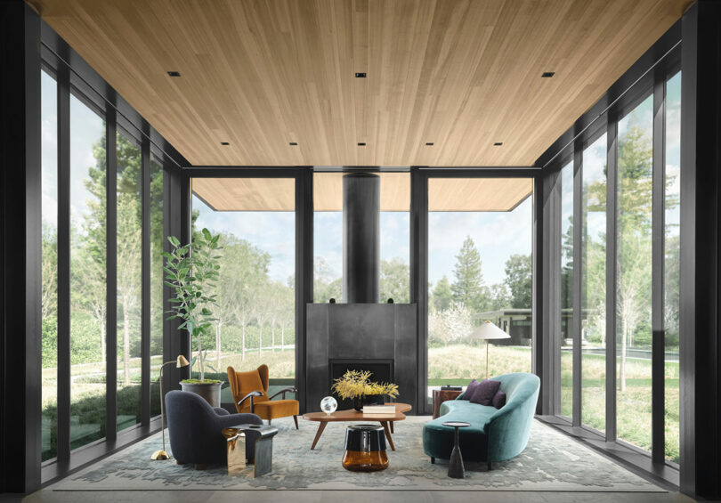 Modern living room with large windows, comfortable seating, a central coffee table, and a fireplace. The room has a natural view and a wooden ceiling.