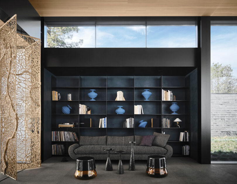 Modern living room with a dark shelving unit displaying blue vases and books. A gray sofa is set in front with two metallic side tables. A decorative screen is to the left. Large windows above.