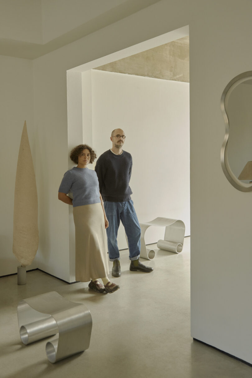 Two people stand in a minimalist room with modern furniture and sculptures from the Alu Archive for Space, featuring neutral tones and geometric shapes.