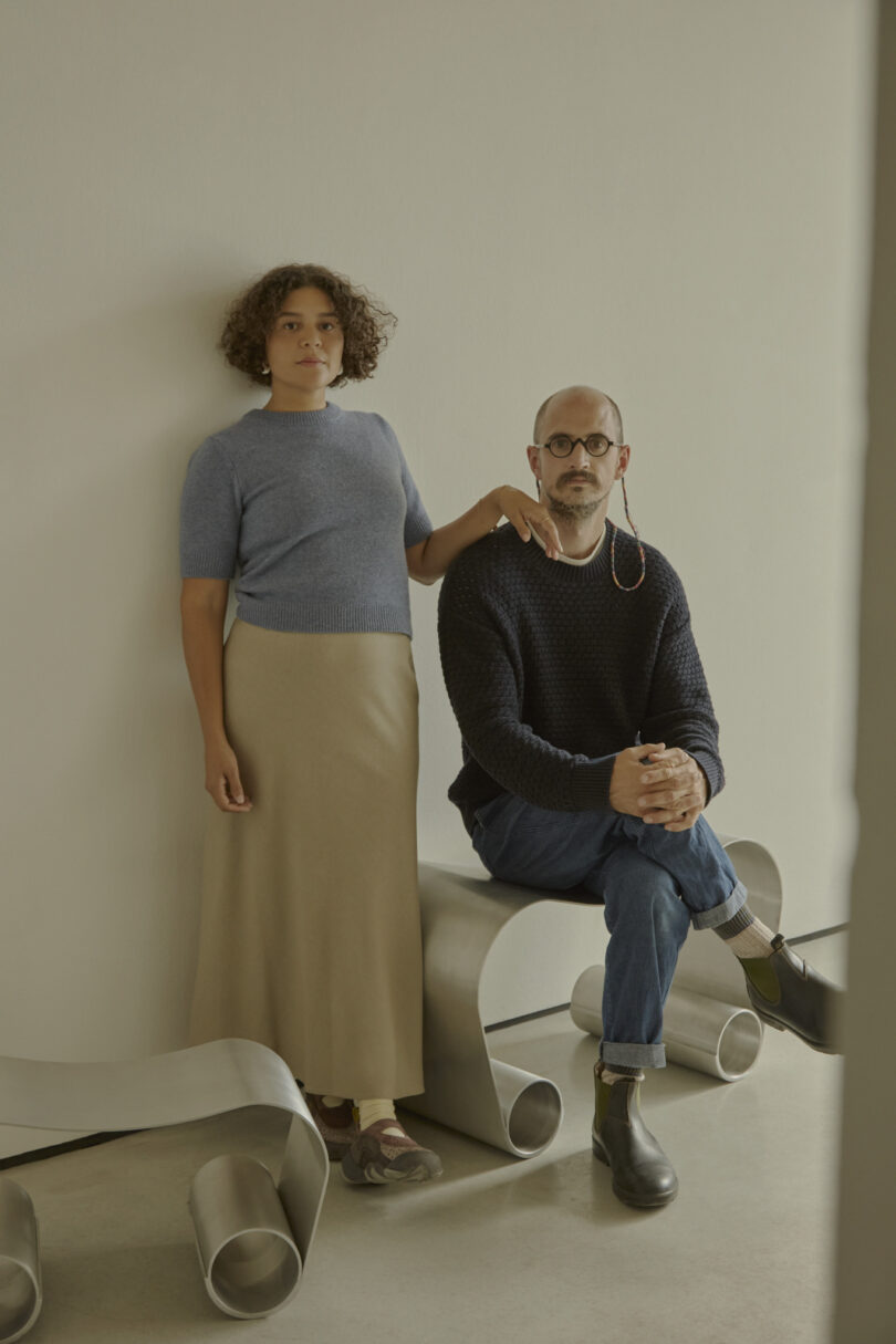 Two people pose indoors, one standing and one seated on a curved metallic structure reminiscent of an Alu Archive for Space. The person standing dons a blue top and beige skirt, while the seated person is casually dressed in a dark sweater and jeans.