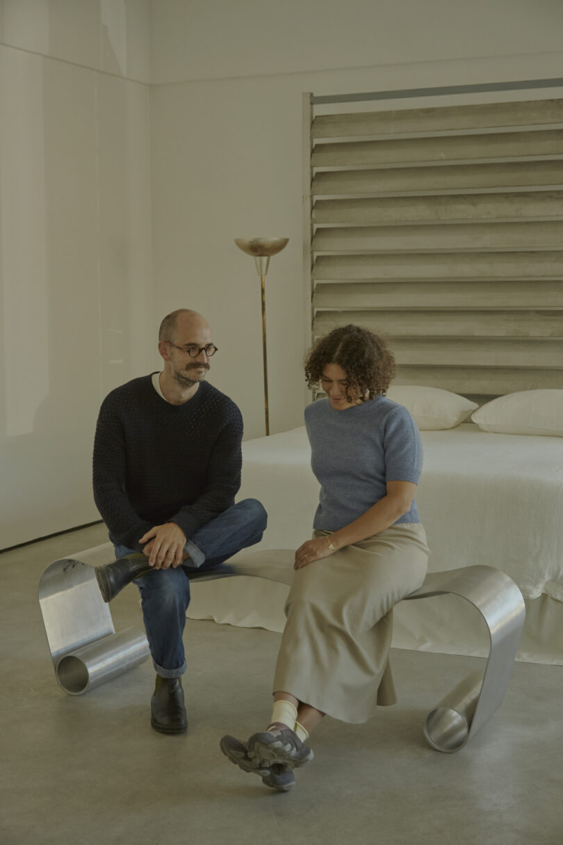 A man and woman sit on a metallic bench in a minimalist room, an Alu Archive for Space, with a bed and a tall lamp.