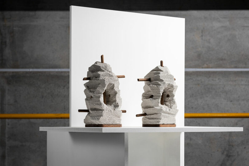 Two abstract stone sculptures with wooden rods on a white pedestal and a white backdrop, against a gray concrete wall with yellow piping.