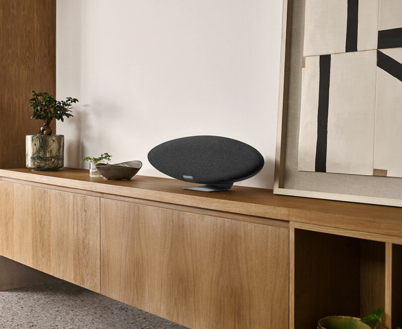 A modern Zeppelin Pro Edition speaker sits on a wooden sideboard with a plant and a painting nearby.