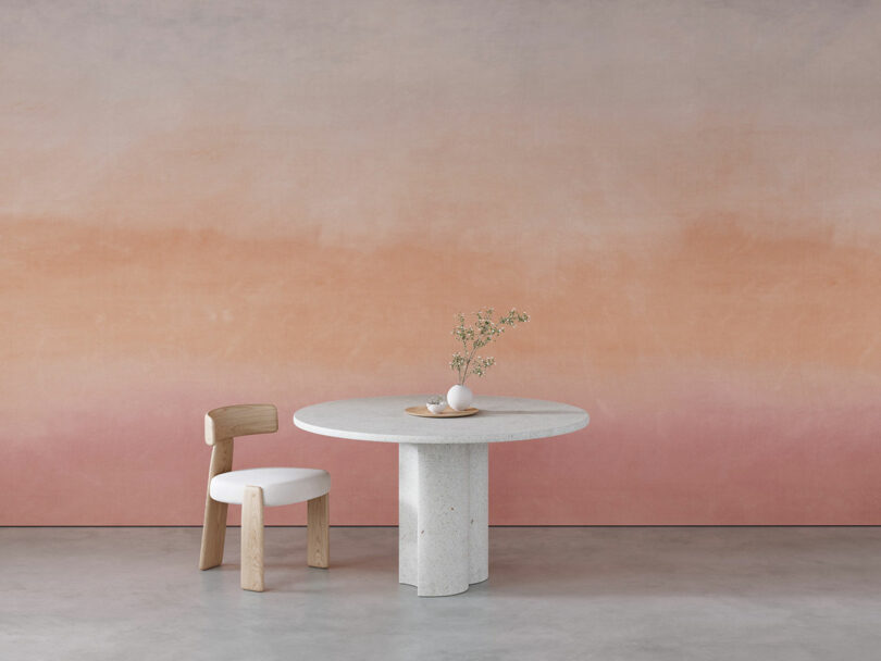 A minimalist setup with a round stone table and a wooden chair on a concrete floor, against a gradient peach to pink wall. A small vase with flowers and a bowl are on the table.