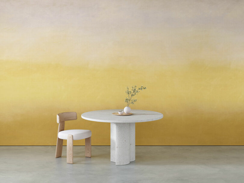 Minimalist setting with a round stone table, a wooden chair with a white cushion and a simple vase with flowers. The background is a gentle gradient from yellow to beige.