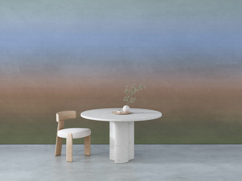 A minimalist setting with a round stone table and chair on a concrete floor. The wall has a color gradient from blue to brown. There is a small vase with green plants on the table.