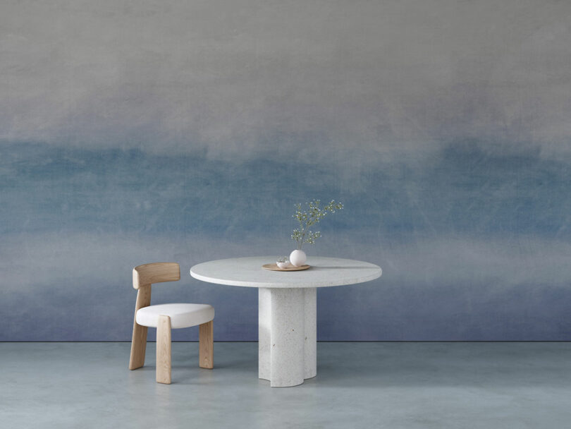 The minimalist room features a round table with a small vase and eggs on it and a wooden chair. The walls have a blue gradient design.