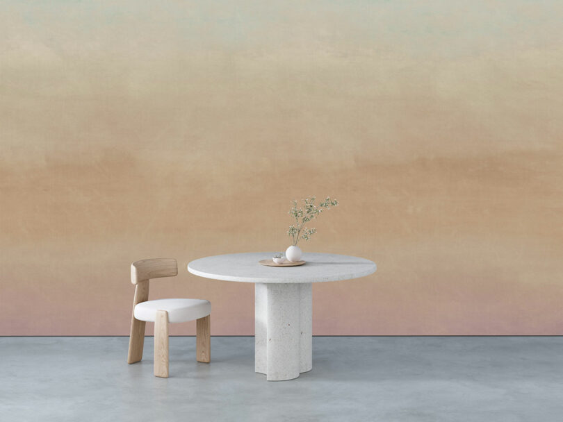 Minimalist setting with a round stone table, a wooden chair with a white cushion, and small vases with branches against a gradient beige wall.