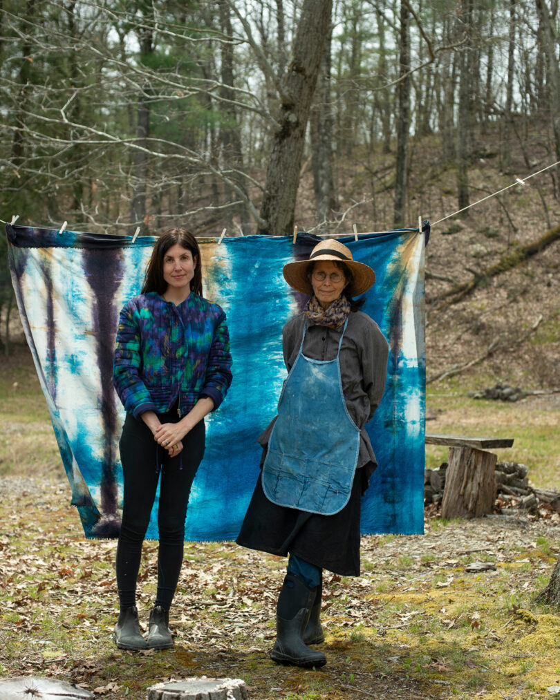 Two people stand outside in front of a piece of batik hanging on a line. The person on the left is wearing a colorful sweater; The person on the right is wearing a hat and apron. Trees and grass are in the background.