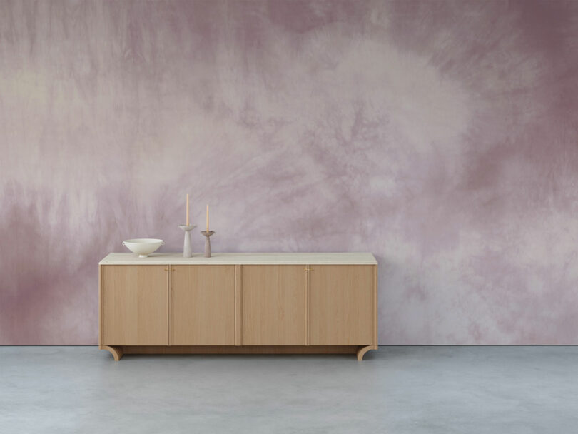 A wooden sideboard in front of a pastel purple batik wall with a bowl, two candlesticks and burning candles on it.