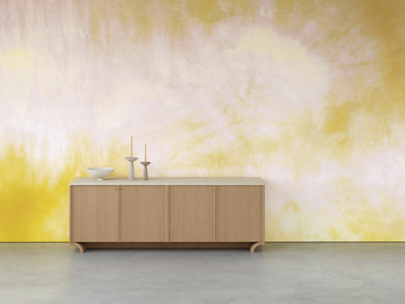 A wooden sideboard with decorative items is placed against a yellow and white abstract wall.