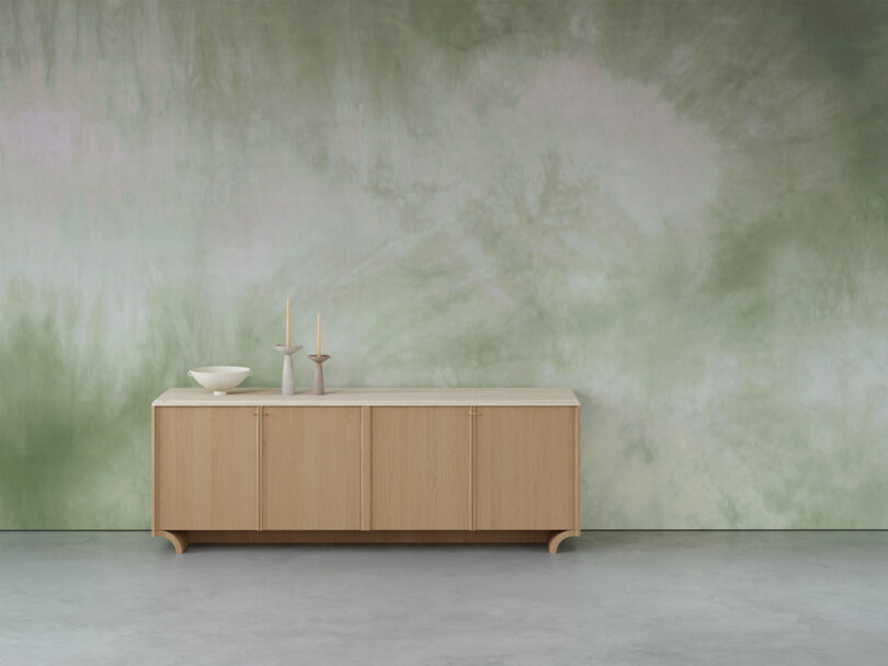 Wooden sideboard with a bowl and two candlesticks on top, set against a green tie-dye patterned wall.