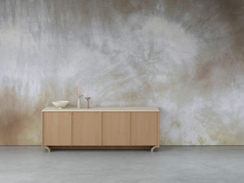 Wooden sideboard with a bowl and two candlesticks, set against a muted, tie-dye patterned wall.