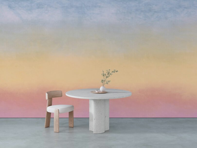 A round, minimalist table with a vase sits on a concrete floor against a gradient wall in pastel blue, yellow, and pink. A wooden chair is beside the table.