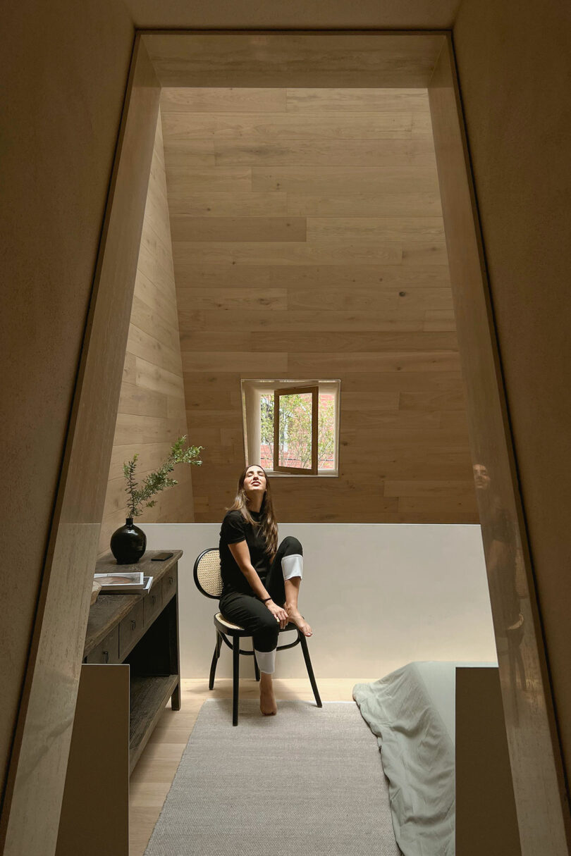 Person sitting on a chair in a small, wooden room with a vaulted ceiling, next to a window and a vase with branches on a table.