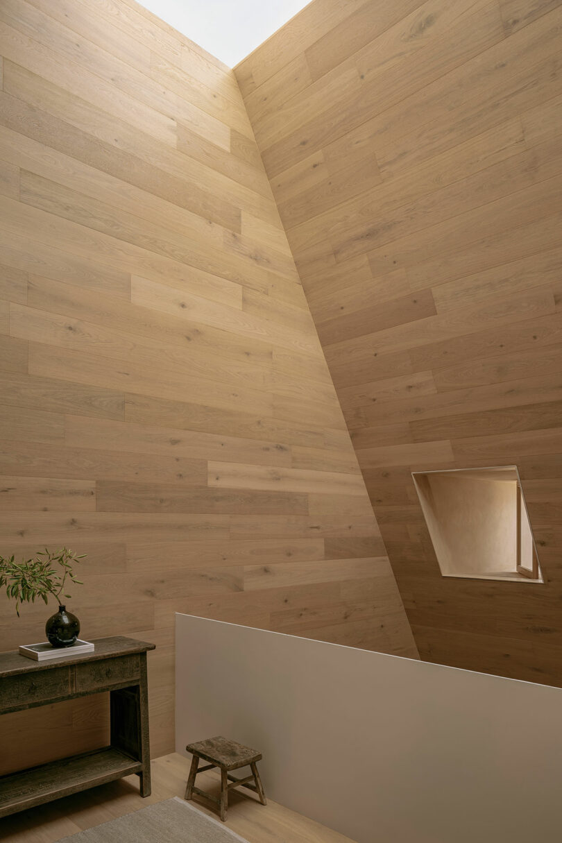 A minimalist interior with wood-paneled walls, a small window, a wooden table with a plant, and a stool. Natural light illuminates the space.