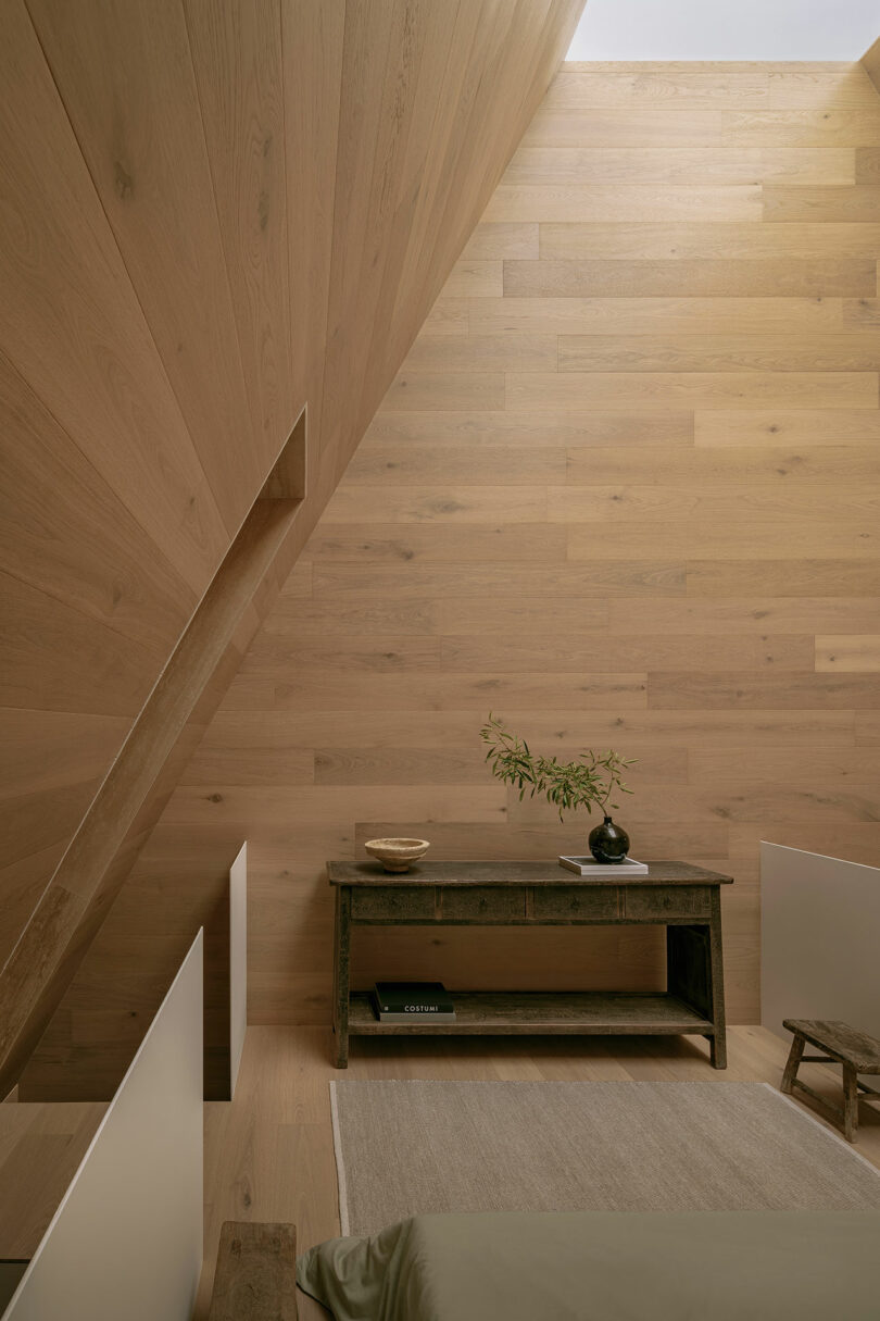 Minimalist wooden interior with a rustic table, small plant, and low bed. Light wood panel walls and ceiling create a warm, natural ambiance.