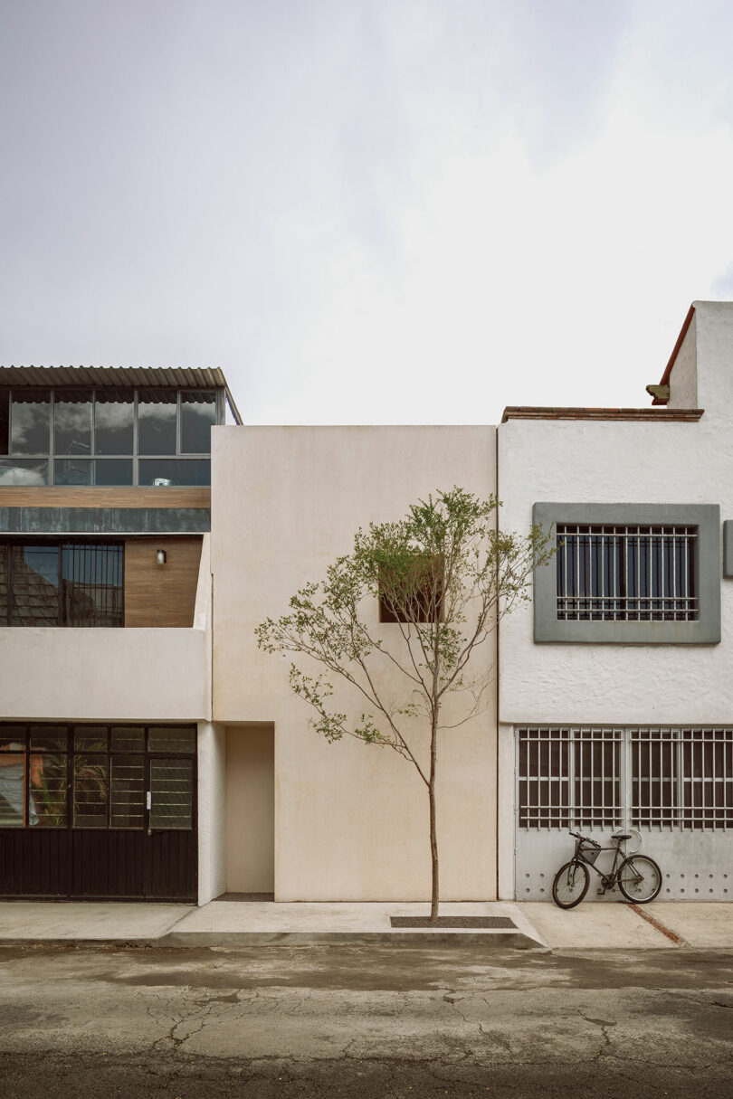 Modern, minimalist urban building with a small tree in front and a bicycle leaning against the adjacent structure, under a cloudy sky.