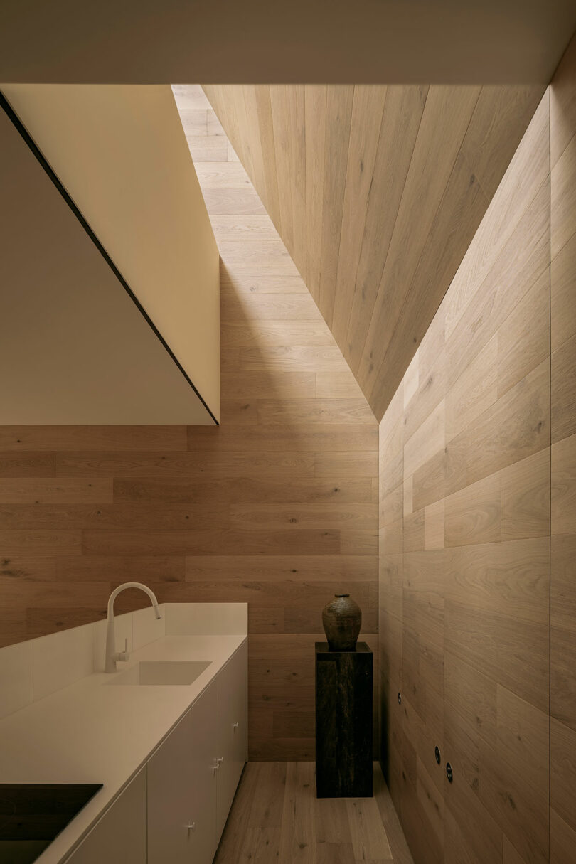 Minimalist kitchen with wood-paneled walls and ceiling, white countertop, sink with faucet, and a small sculpture on a black pedestal. Natural light filters through an angular skylight.