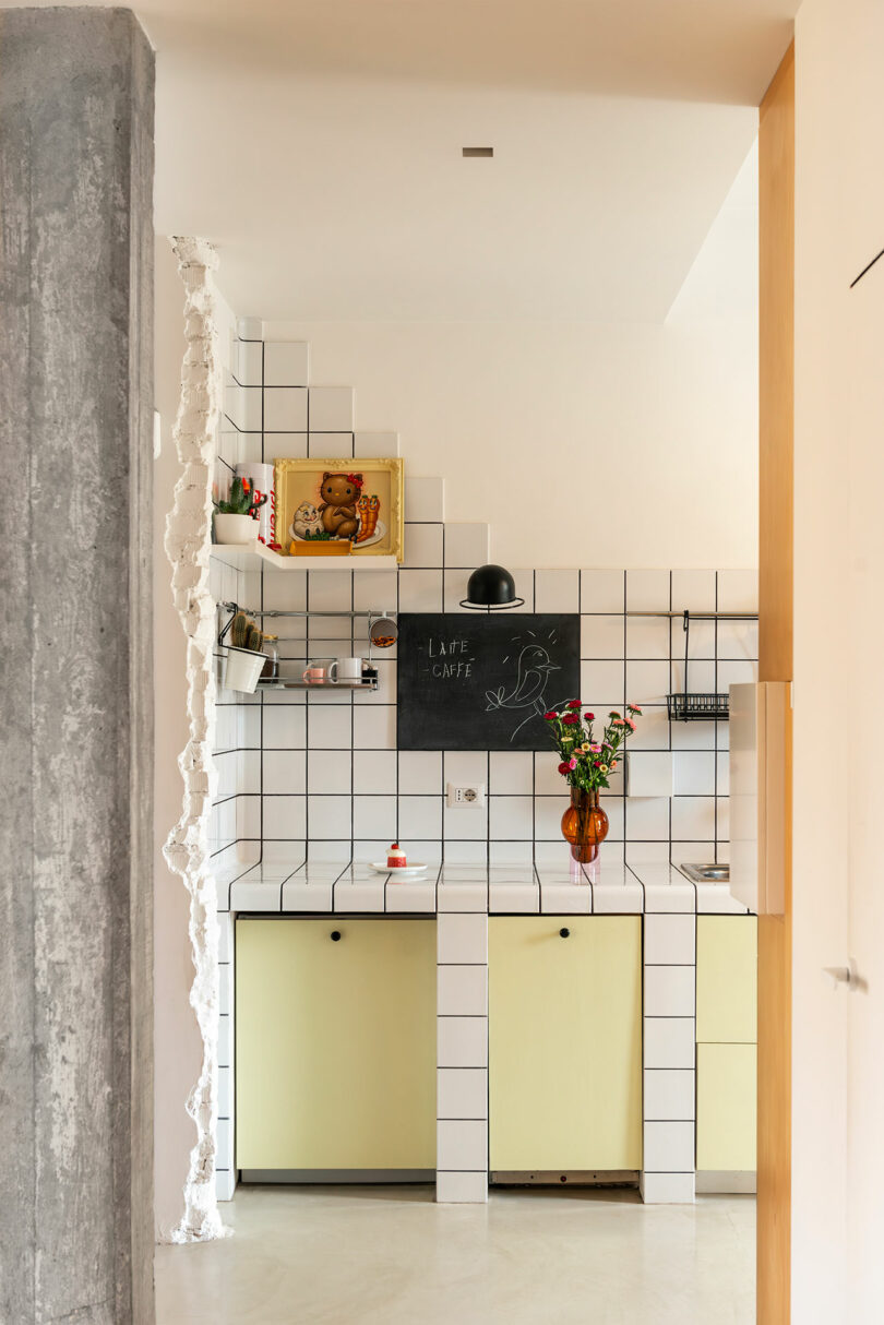 A kitchen with yellow cabinets, white tiled walls, a black chalkboard, and shelves holding décor and plants. A concrete pillar with an exposed edge is visible on the left.