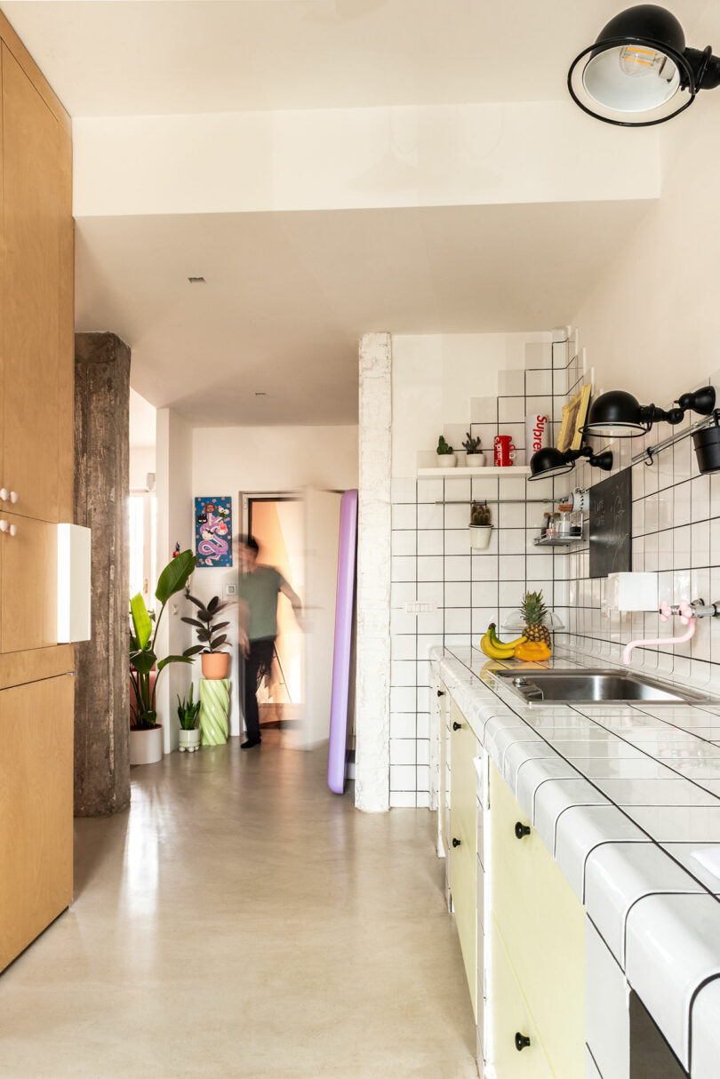 A person walking in a modern kitchen with tiled counters, a sink, shelves with various items, potted plants, and a natural wood partition.