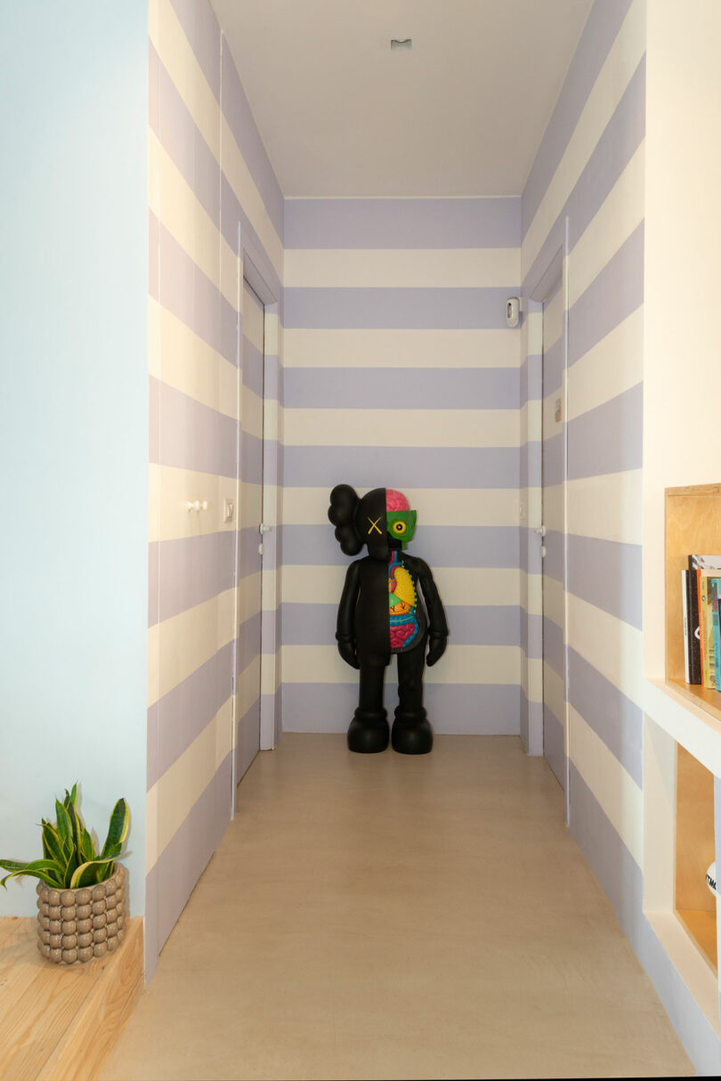 A hallway with wide horizontal striped walls features a large black sculptural figure standing in the center. A plant is on the left, and shelves with books are on the right.