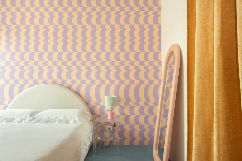 A bedroom features a white bed, a translucent side table with a lamp, a pink-framed mirror, orange curtains, and a wall with a purple and peach geometric pattern.