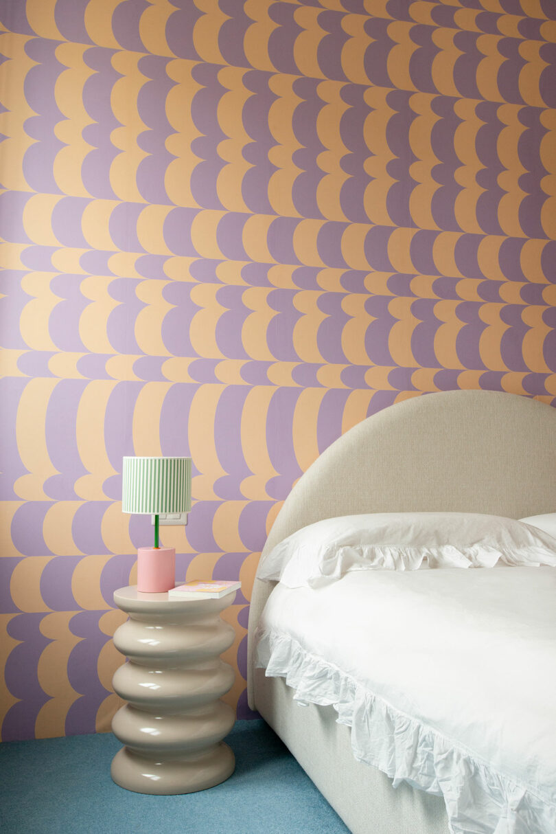 A bedroom with a bed, ruffled white bedding, pastel wavy-patterned wallpaper, and a round side table holding a small green-striped lamp and a pink cup.