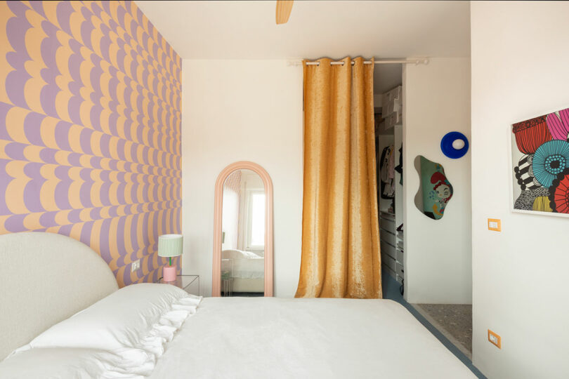 A bedroom with a white bed, pink and yellow patterned accent wall, gold curtain, and eclectic wall art. A small pink mirror and lamp are beside the bed.