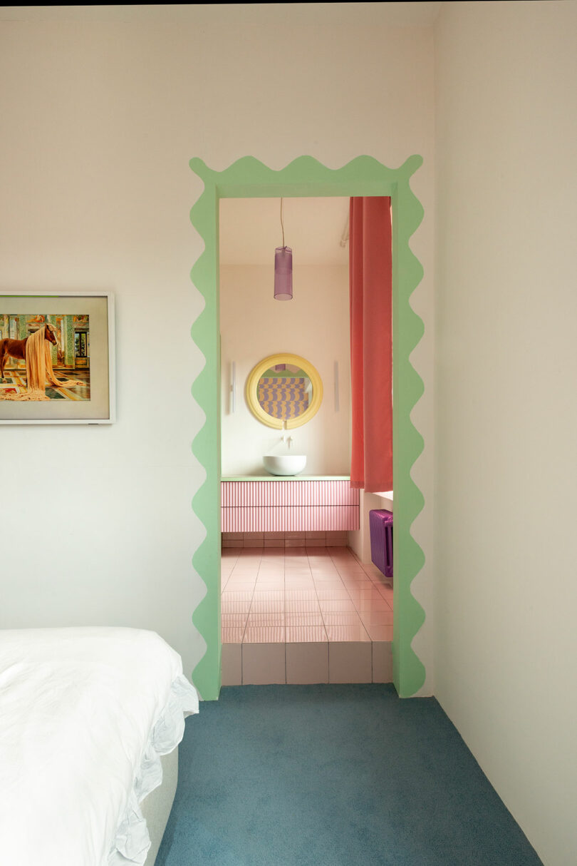 A view through a wavy green doorway shows a room with pastel pink tiles, a circular mirror, and a purple lamp. A framed picture hangs on the left wall.