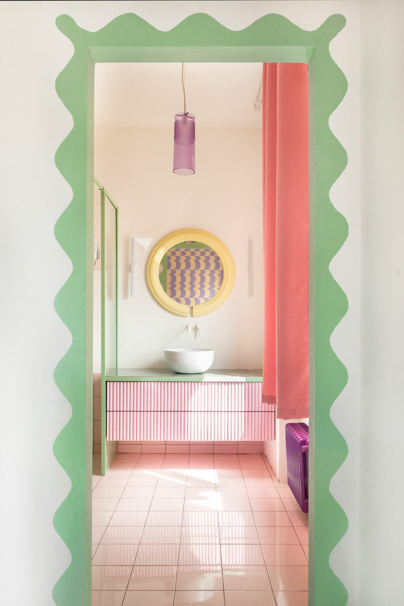 View of a colorful bathroom with a green scalloped doorway. Features a pink vanity, round mirror, and purple pendant light. Walls and floor are light-colored with pink curtain accents.