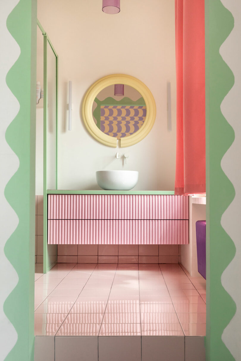 Colorful bathroom with a round mirror, pastel pink striped vanity, vessel sink, and green and pink decor elements.