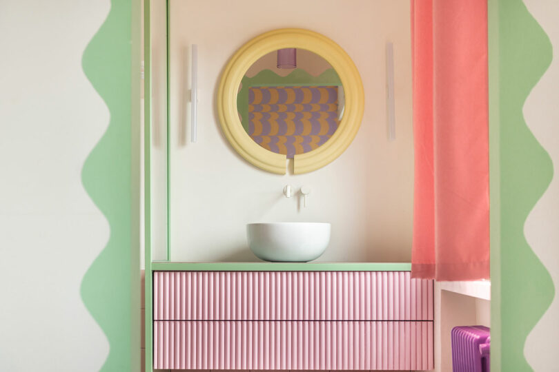 Pastel-themed bathroom with a round mirror, a white vessel sink, and a pink-striped vanity. Green and pink curtains frame the space.