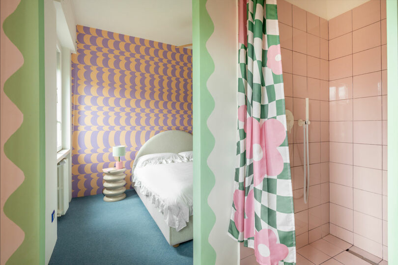 Room with a pastel color scheme, featuring a bed, a patterned accent wall, and a bathroom with pink tiles and a colorful shower curtain.