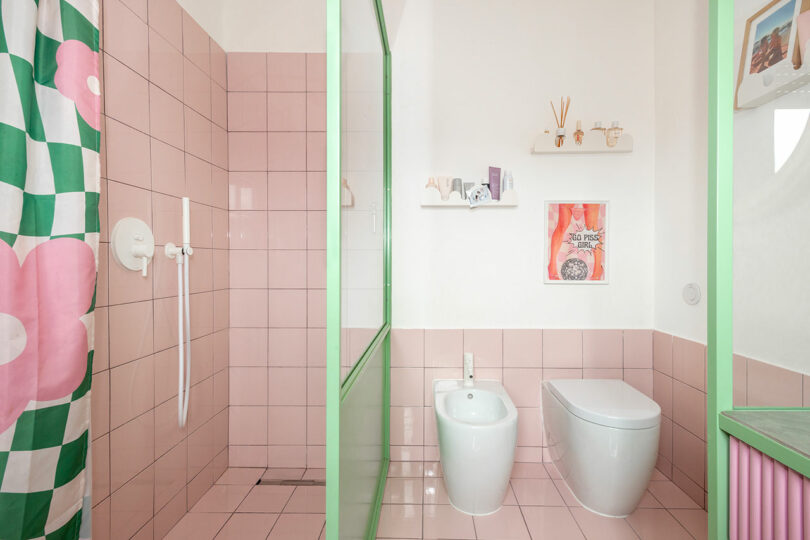 A bathroom with pink tiles, a shower with a floral curtain, and a toilet and bidet. A green frame separates the shower area, and a shelf with decor is above the toilet.