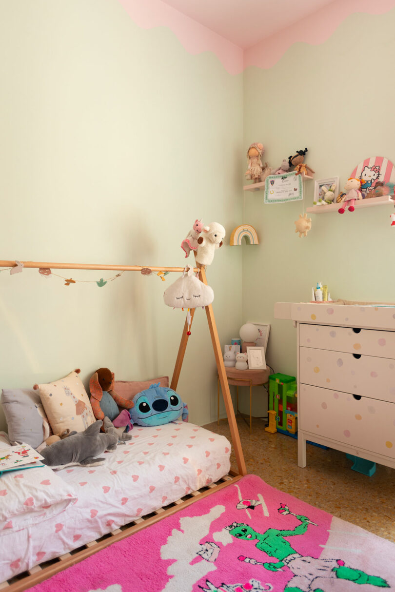 A pastel-themed children's room with a floor bed, soft toys, a colorful dresser, and wall shelves holding books and decor items. A pink rug with green patterns lies on the floor.