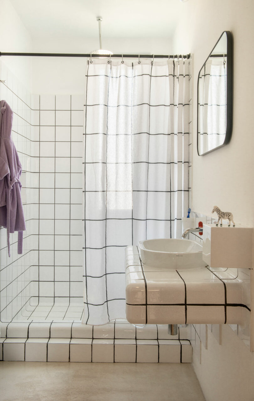 A minimalist bathroom with a walk-in shower, black and white grid tiles, a white shower curtain, a round basin on a tiled countertop, a wall mirror, and a purple robe hanging on the wall.