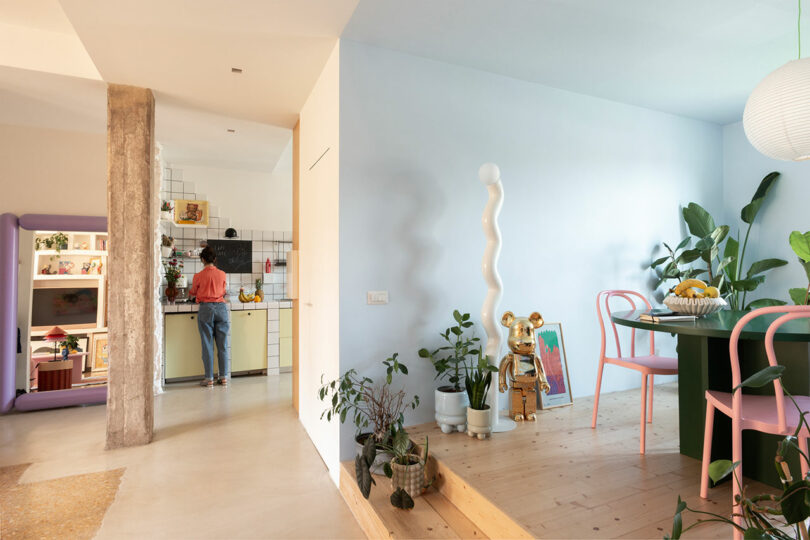 A person is cooking in a colorful kitchen with plants and modern decor, including a green dining table, pink chairs, and a tall white sculpture.